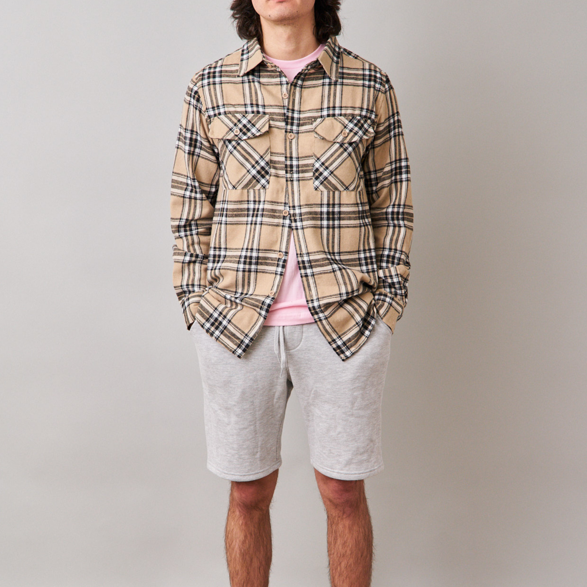 man wear beige long sleeve flannel with pink tshirt underneath. He is also wearing gray jogger short with a drawstring waistband. 