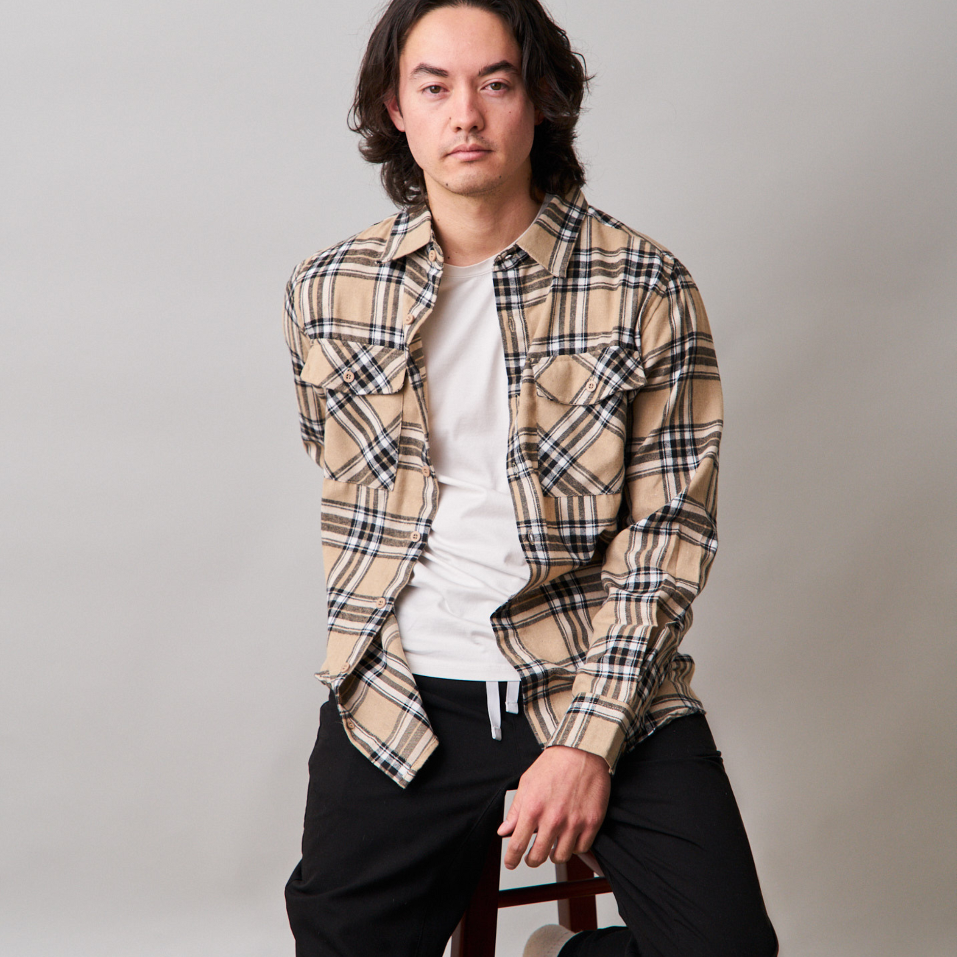 man is sitting on stool wearing long sleeve beige flannel shirt, white tshirt with black sweatpants.